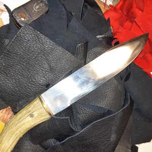 May include: A close-up of a knife with a wooden handle and a silver blade. The knife is lying on a pile of black leather scraps.