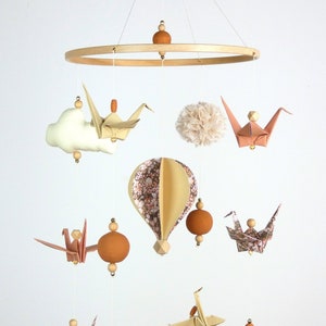 May include: A wooden mobile with a circular frame, featuring a variety of hanging elements, including origami cranes, clouds, pom-poms, and a hot air balloon. The mobile is decorated with a floral pattern and has a natural wood finish.