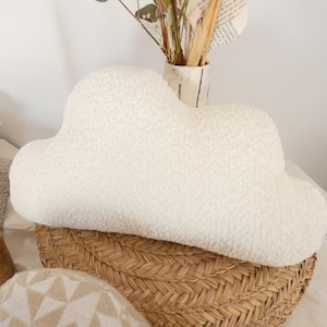 Peut inclure: Un coussin en forme de nuage, de couleur blanc cassé et à la texture duveteuse. Il est posé sur un panier en osier. D'autres coussins et objets décoratifs sont visibles en arrière-plan.