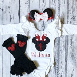 May include: A white long-sleeved bodysuit with a black and red glitter Minnie Mouse design, the number 1, and the name "Milania" embroidered on the front. The outfit includes a red and white polka dot headband with black mouse ears and black leg warmers with red glitter Mickey Mouse ears.