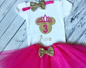 Può includere: Una maglietta bianca con una testa di Topolino dorata con glitter, una corona e il numero 3 in glitter rosa. La maglietta ha anche il nome Ava scritto in glitter dorato. La maglietta è abbinata a una gonna di tulle rosa e un fiocco di glitter dorato.