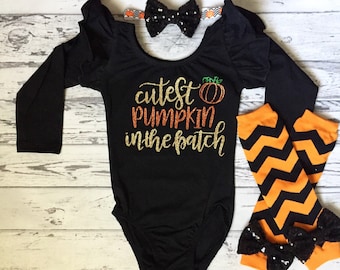 Może przedstawiać: Czarny, długorękawny body z napisem "cutest pumpkin in the patch" w złotym brokacie i małym pomarańczowym nadrukiem dyni. Body ma falbankę przy dekolcie i jest sprzedawany z czarną i białą opaską z kokardką oraz pomarańczowo-czarnymi legginsami w chevron z czarną kokardką z cekinów.