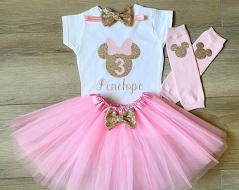 Può includere: Un tutù rosa con un fiocco dorato, una maglietta bianca con una testa di Minnie Mouse con glitter dorati e il numero 3, e scaldagambe rosa con orecchie di Topolino con glitter dorati. La maglietta dice anche "Penelope".