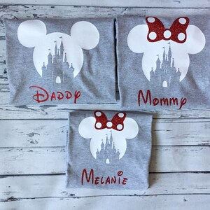 May include: Three grey t-shirts with a white silhouette of Mickey Mouse ears and Cinderella's castle. The shirts have red glitter text that says "Daddy", "Mommy", and "Melanie".