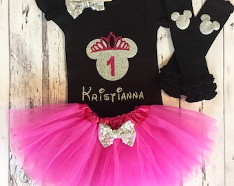 May include: A black t-shirt with a silver glitter Minnie Mouse head with a pink crown and the number 1. The shirt also has the name "Kristianna" in gold glitter. The shirt is paired with a pink tulle tutu and black arm warmers with silver glitter Mickey Mouse ears.