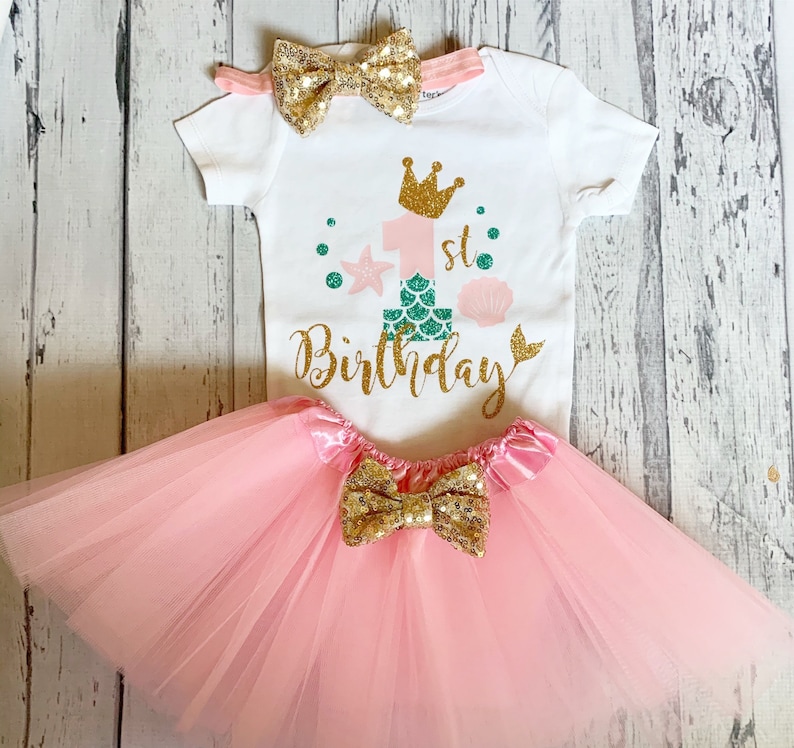 May include: A white onesie with a pink and green mermaid-themed design that says "1st Birthday". The onesie is paired with a pink tulle tutu and a gold glitter bow.