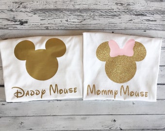 May include: Two white t-shirts with gold glitter Mickey Mouse silhouettes. The left shirt says "Daddy Mouse" and the right shirt says "Mommy Mouse".