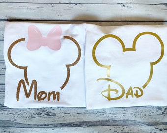 May include: Two white t-shirts with gold glitter Mickey Mouse head outlines. The shirt on the left has a pink bow on top of the head and the word "Mom" underneath. The shirt on the right has the word "Dad" underneath.
