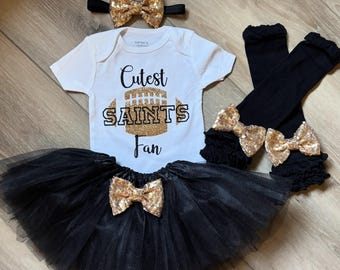 Puede incluir: Conjunto de ropa para bebé que incluye un body blanco con el texto "Cutest Saints Fan", un tutú negro, calentadores y una diadema. Cada artículo está adornado con lazos de lentejuelas doradas. El conjunto es para un bebé.
