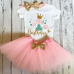 May include: A pink tulle tutu with a gold glitter bow, a white onesie with a gold glitter crown, the number one with a mermaid tail, and the text "1st Birthday", and a gold glitter bow headband.
