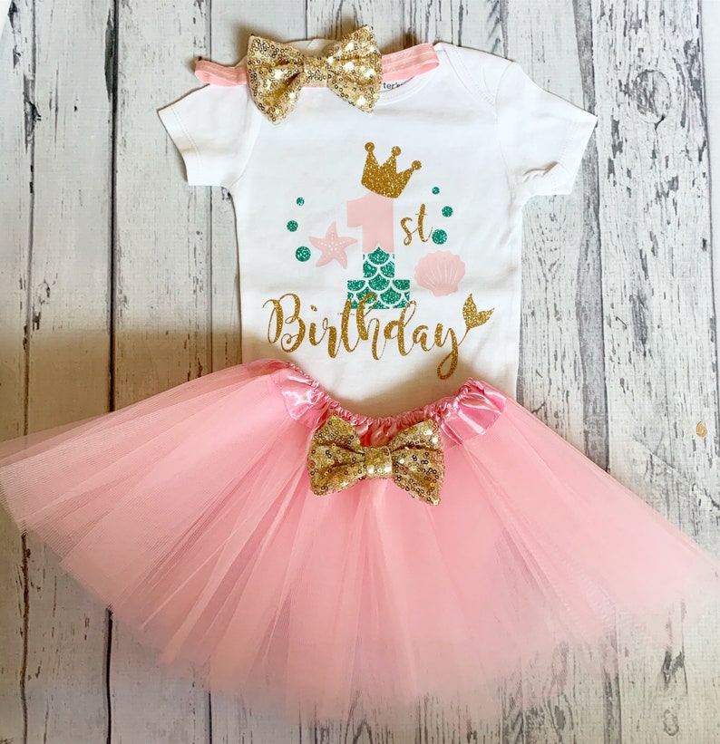 May include: A pink tulle tutu with a gold glitter bow, a white onesie with a gold glitter crown, the number one with a mermaid tail, and the text "1st Birthday", and a gold glitter bow headband.