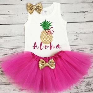 May include: A white tank top with a gold glitter pineapple and the word "Aloha" in pink glitter. The tank top is paired with a pink tulle tutu and a gold glitter bow.