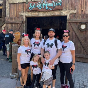 May include: A family of five wearing white t-shirts with a Mickey Mouse silhouette and the words "Mom", "Dad", "Nana", "Grayson", and "Brookelynn" printed on them. They are standing in front of the entrance to Splash Mountain at Disney World.