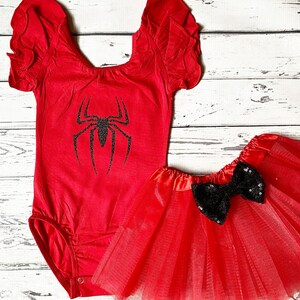 May include: Red and black spider-themed baby outfit. The outfit includes a red leotard with a black spider graphic and a red tulle skirt with a black sequin bow.