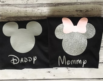 May include: Two black t-shirts with a silver glitter Mickey Mouse head design. One shirt says "Daddy" and the other says "Mommy".