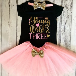 May include: A black t-shirt with the text "Young Wild & Three" in gold glitter with pink hearts and an arrow. The shirt is paired with a pink tulle skirt and a gold glitter bow tie.