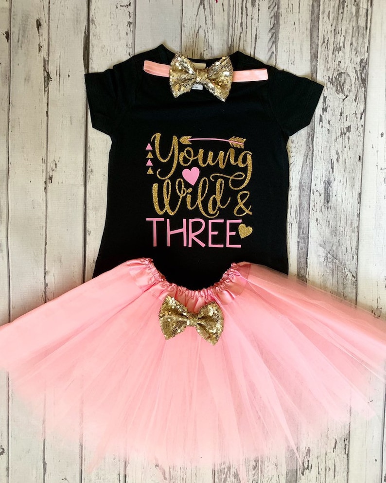 May include: A black t-shirt with the text "Young Wild & Three" in gold glitter with pink hearts and an arrow. The shirt is paired with a pink tulle skirt and a gold glitter bow tie.