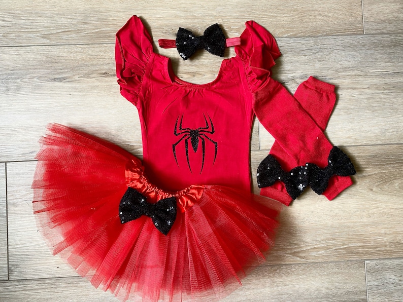 May include: Red and black spider-themed costume set for a child, including a red leotard with a black spider graphic, a red tulle tutu with a black bow, red arm warmers with black bows, and a red headband with a black bow.