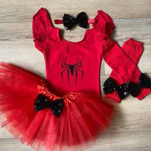 May include: Red and black spider-themed baby girl outfit. The outfit includes a red leotard with a black spider graphic, a red tulle tutu, red leg warmers with black bows, and a black bow headband.