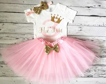 Può includere: Un tutù di tulle rosa con un fiocco dorato, un body bianco con una corona dorata e il numero "3" in rosa con il testo "Me 3rie", e una fascia dorata con un fiocco.