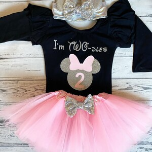 May include: A black long-sleeved shirt with a pink and gold Minnie Mouse design and the text "I'm Two-Dles". The shirt is paired with a pink tulle skirt and a silver headband with ears and a bow.