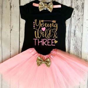 May include: A black t-shirt with the text "Young Wild & Three" in gold glitter with pink hearts and an arrow. The shirt is paired with a pink tulle skirt and a gold glitter bow tie.