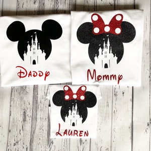 May include: Three white t-shirts with black Mickey Mouse ears and a silhouette of Cinderella's castle. The shirts have the words "Daddy", "Mommy", and "Lauren" printed on them.