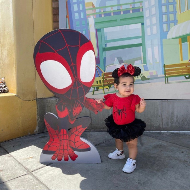 May include: A young child wearing a red and black Spider-Man costume with a black tulle tutu skirt and a red bow headband. The child is standing in front of a large cardboard cutout of Spider-Man.