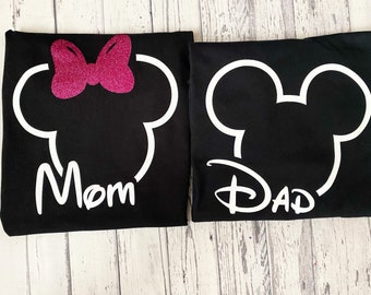 May include: Two black t-shirts with white Mickey Mouse head outlines. One shirt has a pink glitter bow and the word "Mom" in white. The other shirt has the word "Dad" in white with a Disney font.
