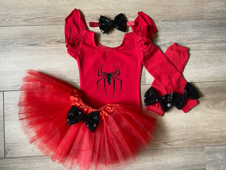 May include: Red and black spider-themed baby girl outfit. The outfit includes a red leotard with a black spider graphic, a red tulle tutu, red leg warmers with black bows, and a black bow headband.