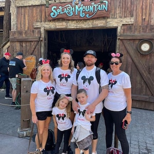 May include: A family of five wearing white t-shirts with a Mickey Mouse silhouette and the words "Mom", "Dad", "Nana", "Grayson", and "Brookelynn" printed on them. They are standing in front of the entrance to Splash Mountain at Disney World.