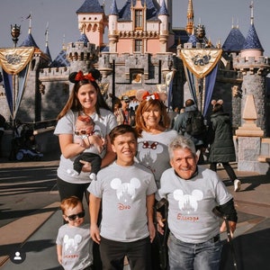 May include: A family of five wearing matching gray t-shirts with a white Mickey Mouse silhouette and the words "Dad", "Grandpa", and "Jackson" printed on them. They are standing in front of Sleeping Beauty's Castle at Disneyland.