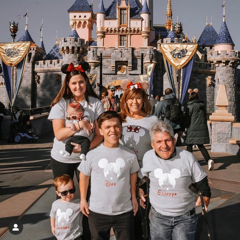 May include: A family of five wearing matching gray t-shirts with a white Mickey Mouse silhouette and the words "Dad", "Grandpa", and "Jackson" printed on them. They are standing in front of Sleeping Beauty's Castle at Disneyland.