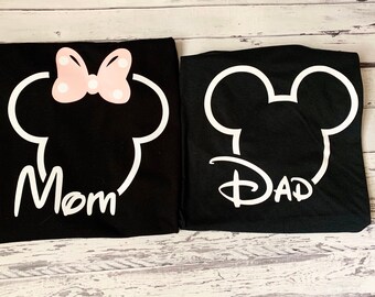 May include: Two black t-shirts with white outlines of Mickey Mouse ears. The left shirt says "Mom" and has a pink bow with white polka dots. The right shirt says "Dad".