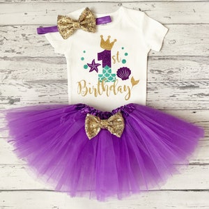 May include: A white onesie with a gold glitter '1st Birthday' design featuring a purple mermaid tail, a crown, and seashells. The onesie is paired with a purple tulle tutu and a gold glitter bow.