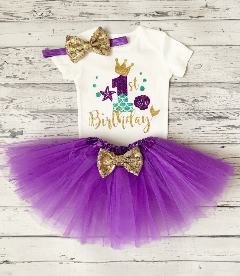 May include: A white onesie with a gold glitter '1st Birthday' design featuring a purple mermaid tail, a crown, and seashells. The onesie is paired with a purple tulle tutu and a gold glitter bow.