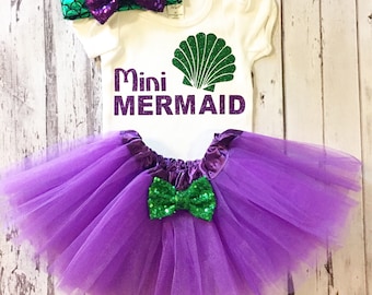 Peut inclure: Un tutu en tulle violet avec un nœud en sequins vert, un body blanc avec un coquillage vert et le texte "Mini Mermaid", et un bandeau en sequins vert et violet.