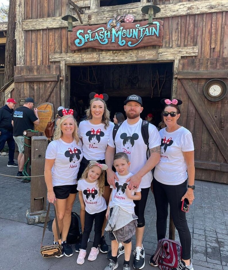 May include: A family of five wearing white t-shirts with a Mickey Mouse silhouette and the words "Mom", "Dad", "Nana", "Grayson", and "Brookelynn" printed on them. They are standing in front of the entrance to Splash Mountain at Disney World.