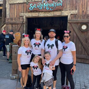 May include: A family of five wearing white t-shirts with a Mickey Mouse silhouette and the words "Mom", "Dad", "Nana", "Grayson", and "Brookelynn" printed on them. They are standing in front of the entrance to Splash Mountain at Disney World.