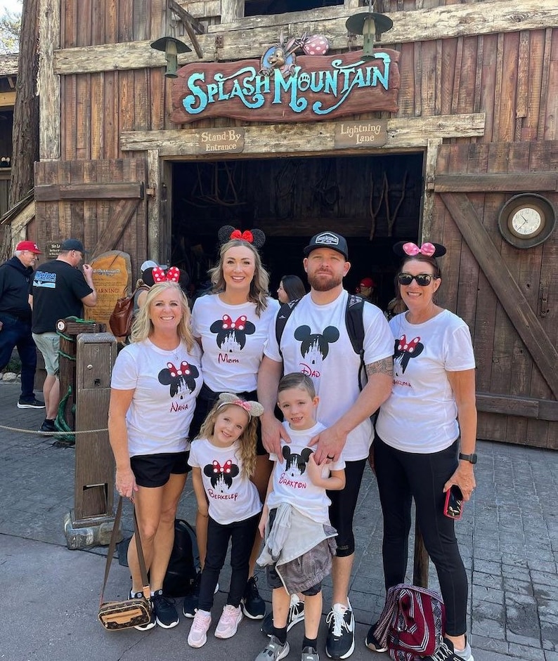 May include: A family of five wearing white t-shirts with a Mickey Mouse silhouette and the words "Mom", "Dad", "Nana", "Grayson", and "Brookelynn" printed on them. They are standing in front of the entrance to Splash Mountain at Disney World.