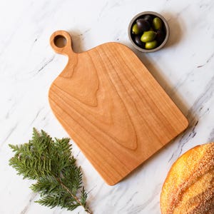 May include: A wooden cutting board with a handle, a small bowl of olives, a loaf of bread, and a sprig of greenery are arranged on a marble surface. The cutting board is light brown and has a smooth finish. The bread is golden brown.