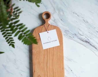 May include: A long, rectangular cherry wood cutting board with a unique handle. The board has a natural wood finish and is labeled "Adirondack Kitchen".