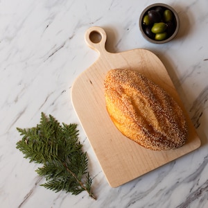 K&ouml;nnte beinhalten: Ein Holzschneidebrett mit Griff h&auml;lt einen Brotlaib mit goldener Kruste. Eine kleine Sch&uuml;ssel mit Oliven und ein Zweig Gr&uuml;nzeug sind auf einer Marmoroberfl&auml;che angeordnet und schaffen eine rustikale Pr&auml;sentation.