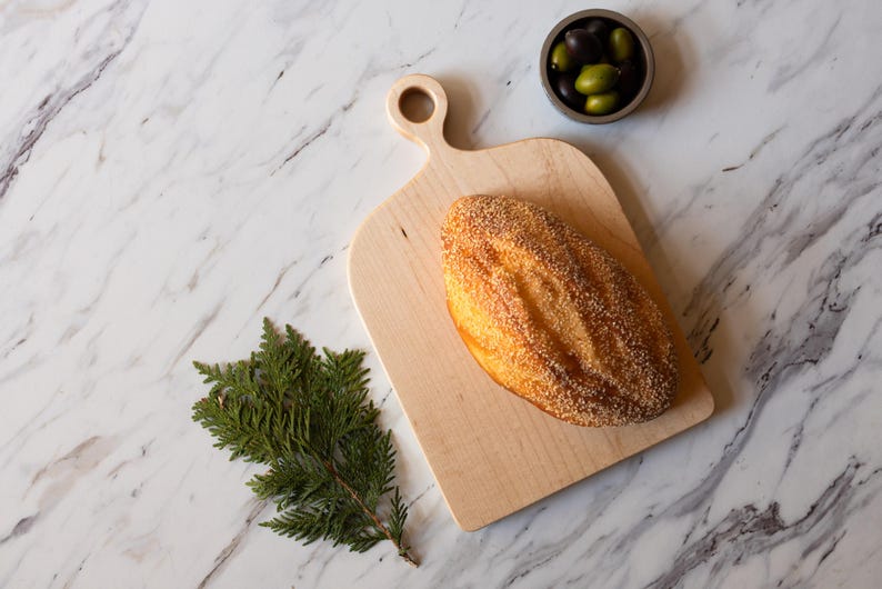 K&ouml;nnte beinhalten: Ein Holzschneidebrett mit Griff h&auml;lt einen Brotlaib mit goldener Kruste. Eine kleine Sch&uuml;ssel mit Oliven und ein Zweig Gr&uuml;nzeug sind auf einer Marmoroberfl&auml;che angeordnet und schaffen eine rustikale Pr&auml;sentation.