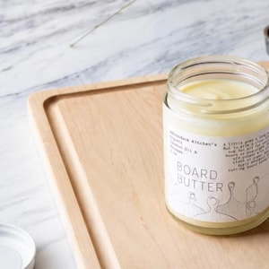 May include: A clear glass jar filled with pale yellow board butter, featuring a white label with the text Adirondack Kitchen's Organic Coconut Oil & Beeswax 3 oz BOARD BUTTER and instructions, sitting on a light-colored wooden cutting board.