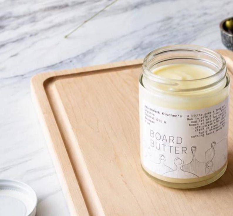 May include: A clear glass jar filled with pale yellow board butter, featuring a white label with the text Adirondack Kitchen's Organic Coconut Oil & Beeswax 3 oz BOARD BUTTER and instructions, sitting on a light-colored wooden cutting board.