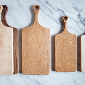 Puede incluir: Cuatro tablas de cortar de madera con asas. Las tablas son de diferentes tama&ntilde;os y est&aacute;n dispuestas en una fila de la m&aacute;s grande a la m&aacute;s peque&ntilde;a. Las tablas est&aacute;n hechas de madera clara y oscura.