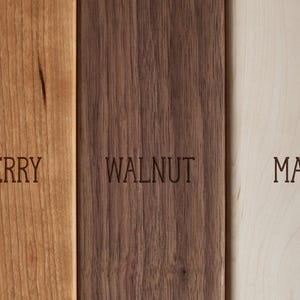 K&ouml;nnte beinhalten: Nahaufnahme von drei Holzbrettern: Kirsche, Walnuss und Ahorn. Das Kirschbrett ist hellbraun, das Walnussbrett dunkelbraun und das Ahornbrett hellbeige. Jedes Brett hat die Holzart in Gro&szlig;buchstaben eingraviert.