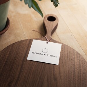 Pode incluir: Uma t&aacute;bua de cortar redonda de madeira com uma al&ccedil;a. A t&aacute;bua &eacute; feita de madeira de noz e tem um acabamento natural. A t&aacute;bua &eacute; etiquetada com "ADIRONDACK KITCHEN" com um logotipo circular.