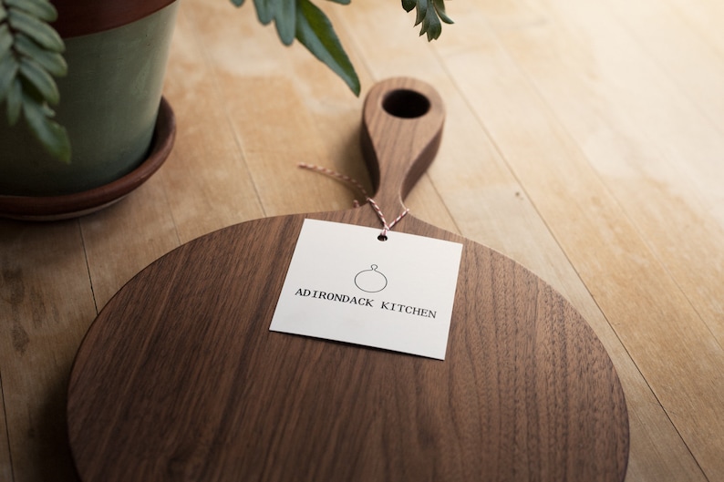 Pode incluir: Uma t&aacute;bua de cortar redonda de madeira com uma al&ccedil;a. A t&aacute;bua &eacute; feita de madeira de noz e tem um acabamento natural. A t&aacute;bua &eacute; etiquetada com "ADIRONDACK KITCHEN" com um logotipo circular.